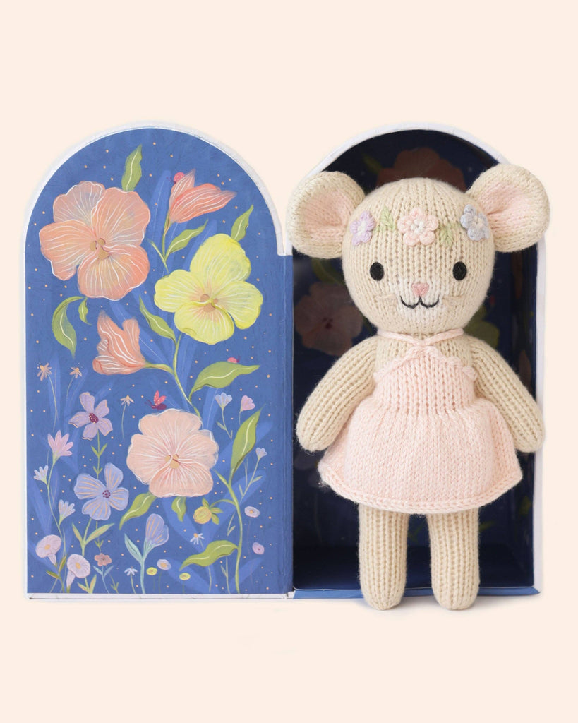 Cuddle + Kind Tiny Emma the Mouse is a hand-knit doll from Peru, dressed in a pink dress and flower headband, presented in an open blue box decorated with colorful painted flowers and stars.