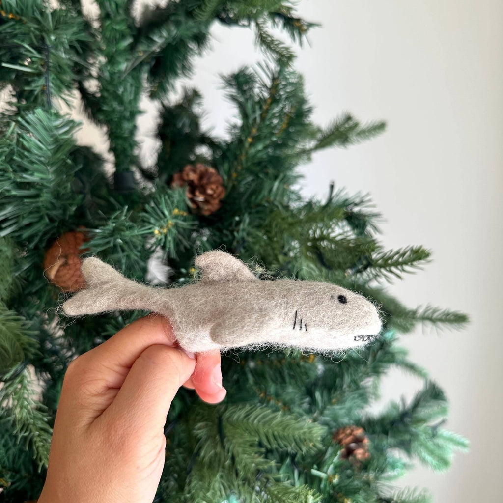 A hand displays the Finger Puppet: Shark, made of gray felt, in front of a Christmas tree with pinecones—perfect for sparking underwater-themed imaginative play.