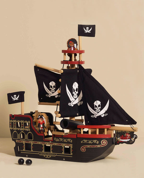 The Barbarossa Pirate Ship & Characters (ships in one week) is an eco-friendly wooden pirate ship toy with black sails, white skull and crossbones, two pirate figures, cannonballs, and a nameplate—shown against a beige background.