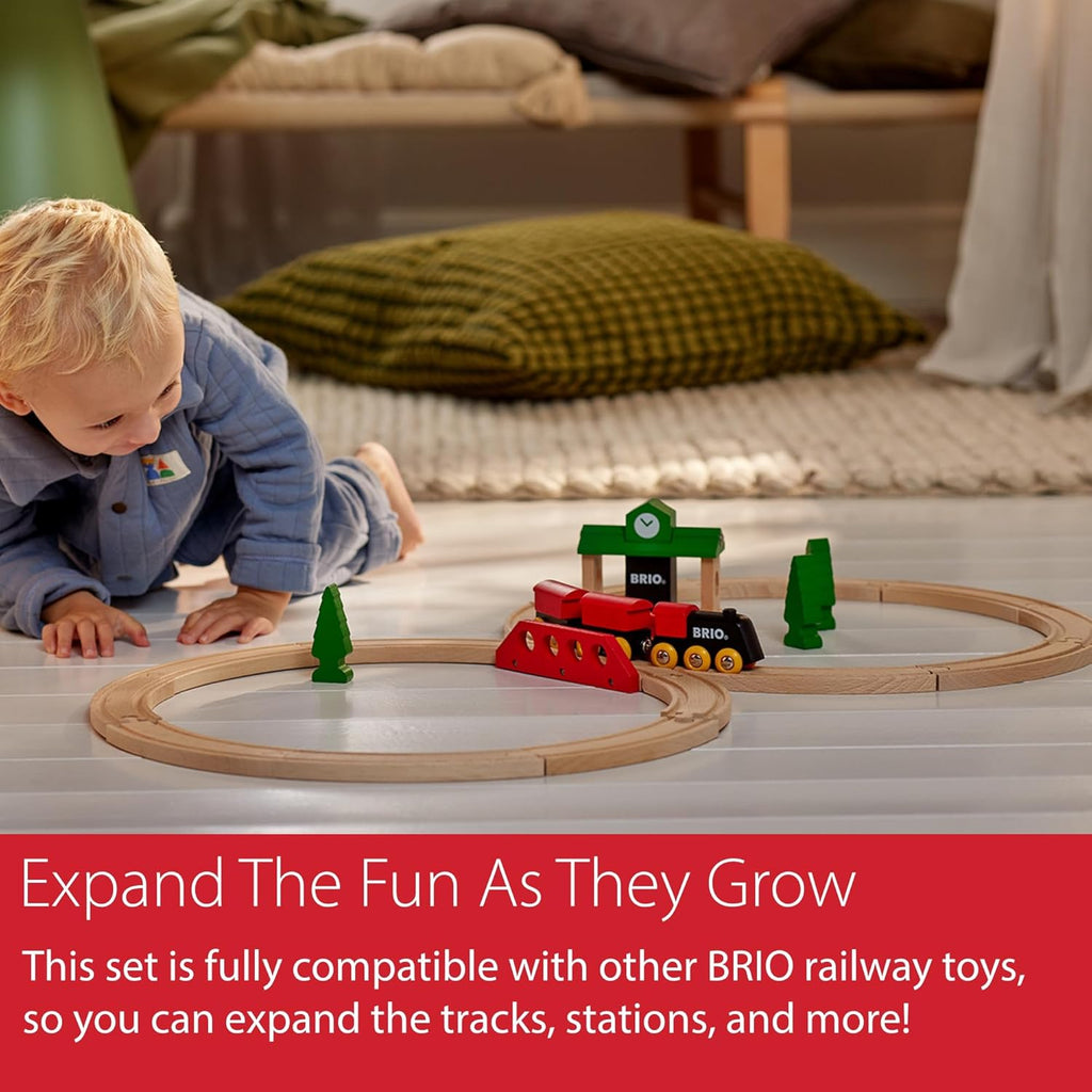 A young child smiles while playing on the floor with the BRIO World Train Set Classic Figure 8 Set, featuring curved tracks, trees, a bridge, and a station. The text highlights how easily this set can be expanded for more fun.