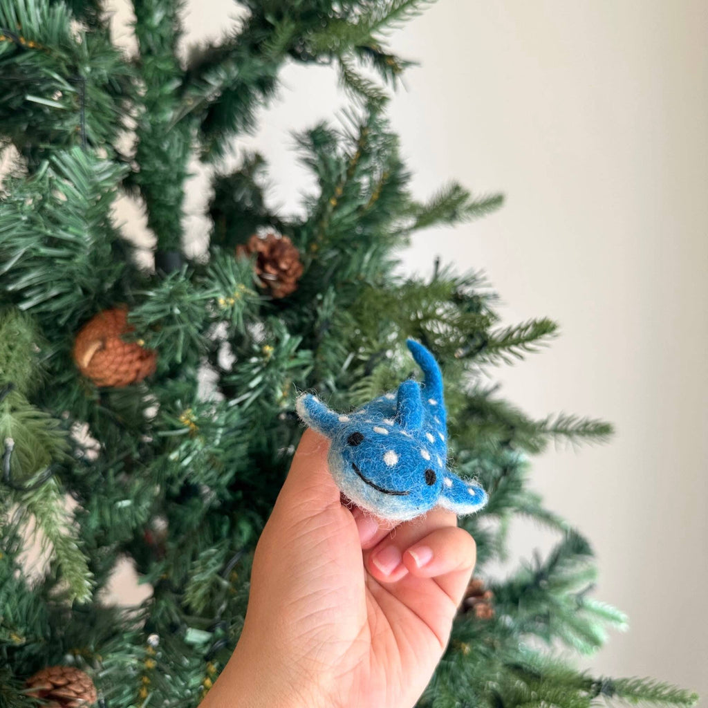 A hand holds a small, blue Finger Puppet: Whale Shark ornament—perfect for imaginative play—in front of a green Christmas tree with pinecones.