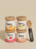 The Yogurt Play Food Pack contains four small containers with wooden lids stacked in two rows. Each container is labeled "Yogurt" and features fruit designs: orange, starfruit, raspberry, and strawberry. A small spoon with a decorative leaf pattern complements this plastic-free set.