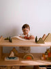 A young child plays with Sabo Concept First Journey—a set of toy cars, mountains, and trees—on a wooden shelf, with colorful wooden toys and blocks neatly arranged against a plain white wall.