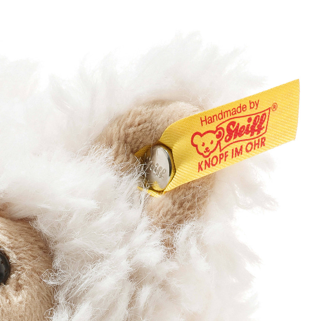 Close-up of a Steiff Leandro Llama Plush Toy, 7 Inches, featuring its ear with a yellow “Handmade by Steiff KNOPF IM OHR” tag and metal button amid soft, fluffy fur—part of the cherished Soft Cuddly Friends collection.