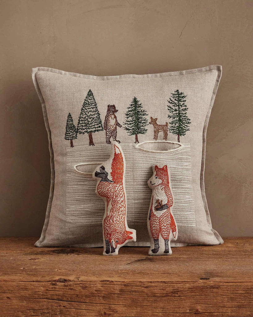 The Coral & Tusk Winter Foxes Pocket Pillow, featuring embroidered trees, a bear, and a deer, rests on wood with two plush fox toys—one upright, one doing a handstand—for charming animal nursery or winter fox decor.