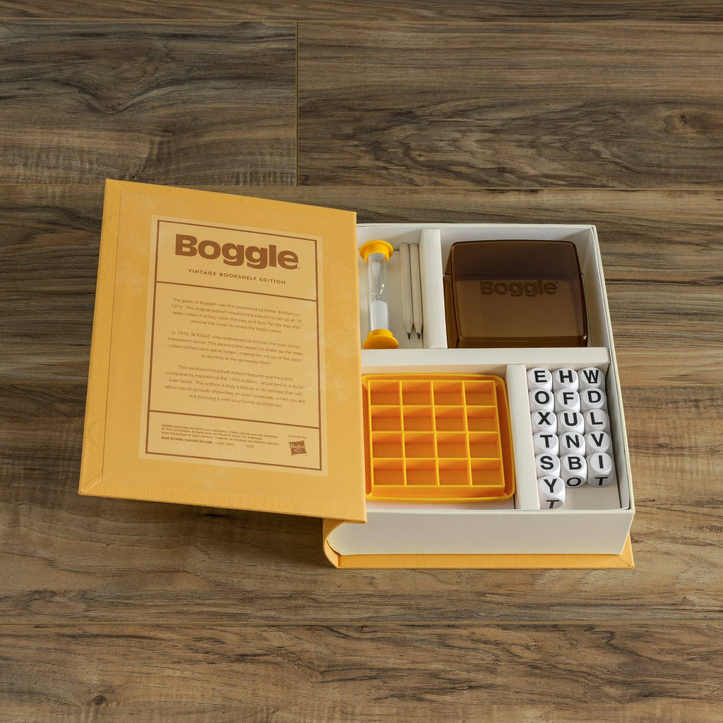 An open Board Game: Boggle Vintage Bookshelf Edition box sits on a wooden surface, revealing the instructions, yellow grid tray, sand timer, brown case, and dice—everything for this classic timed word game.
