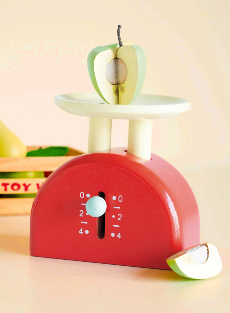 Kitchen Weighing Scales: A red wooden scale with a dial holds a green-and-white apple, with an apple slice beside it and a toy fruit box in the background—perfect for role-play fun in any pretend kitchen.