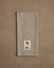 The Coral & Tusk Deck the Halls Bear Tea Towel rests on a textured brown surface, adorned with festive embroidery—ideal as a holiday kitchen towel for gatherings.