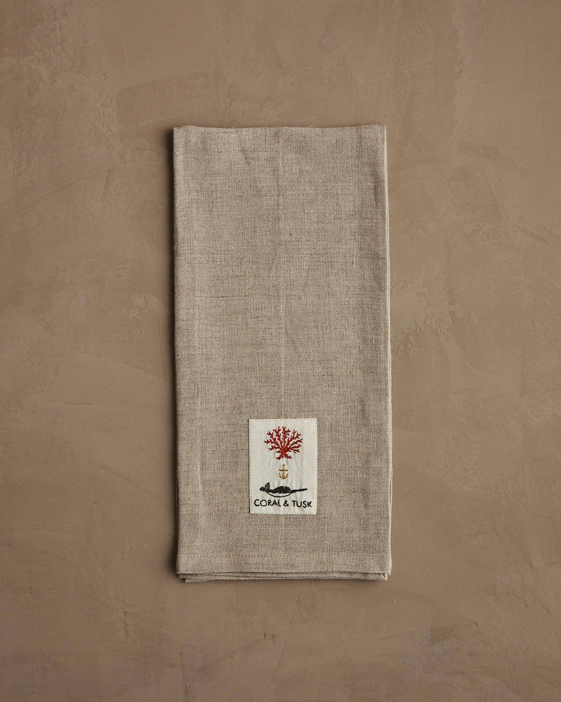 The Coral & Tusk Chipmunks with Pinecone Tea Towel is folded on a taupe textured surface, featuring detailed embroidery near the bottom and the words "CORAL & TUSK" underneath.