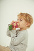 A young child with blond hair wearing a gray long-sleeve shirt holds and pretends to eat Betty The Beetroot Baby Car, a colorful FSC-certified wood toy.