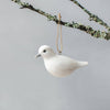 The Hand Carved Hanging Ornament - Dove Bird - 2.5"H hangs by a string from a lichen-covered branch against a plain gray background, offering sustainable, handcrafted decor for any space.