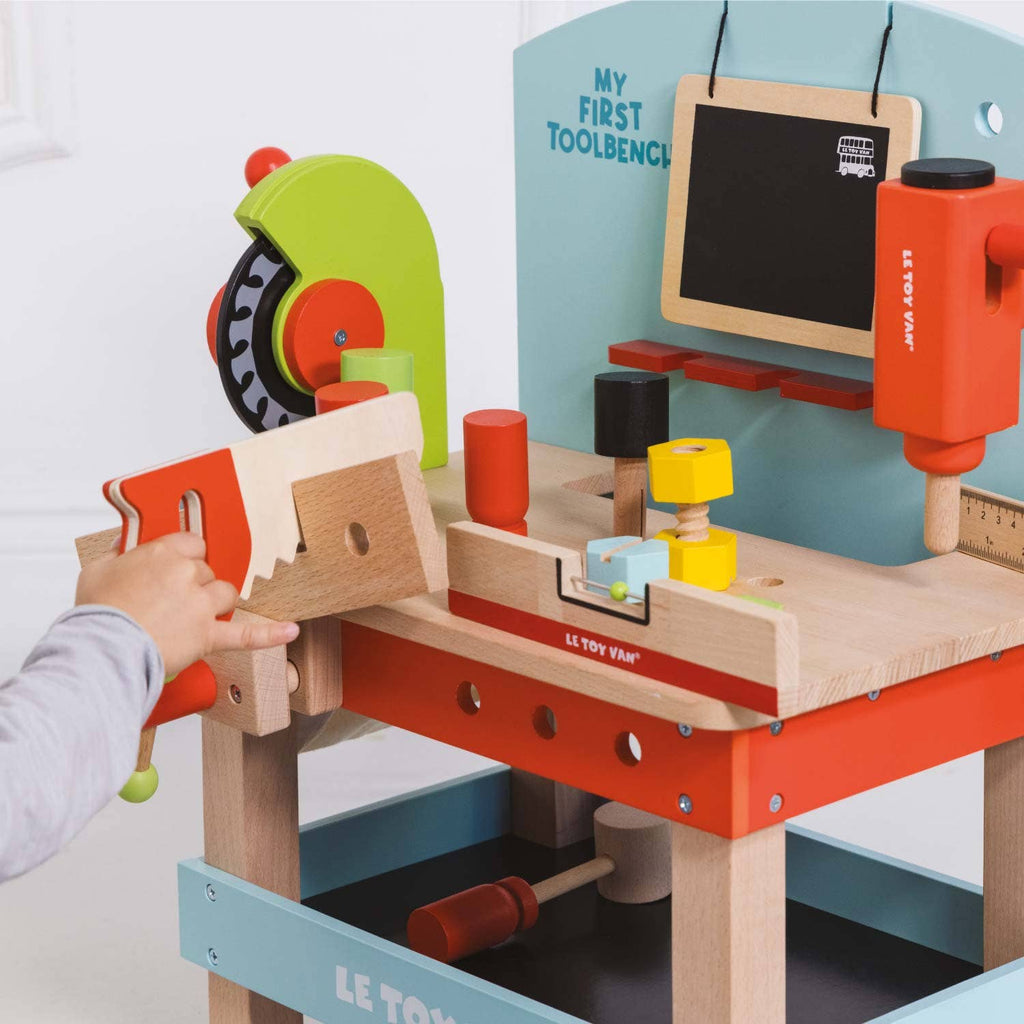 A child uses the Wooden Tool Bench, a colorful playset with a saw, wrench, hammer, nuts, bolts, gears, and a chalkboard labeled "MY FIRST TOOLBENCH," designed to encourage imaginative pretend play.