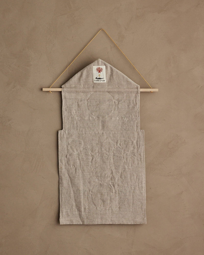The Coral & Tusk Christmas Clock Advent Calendar is a beige fabric wall hanging with pockets, displayed on a wooden rod and string, featuring a small white tag with a red tree design at the top against a tan textured wall.