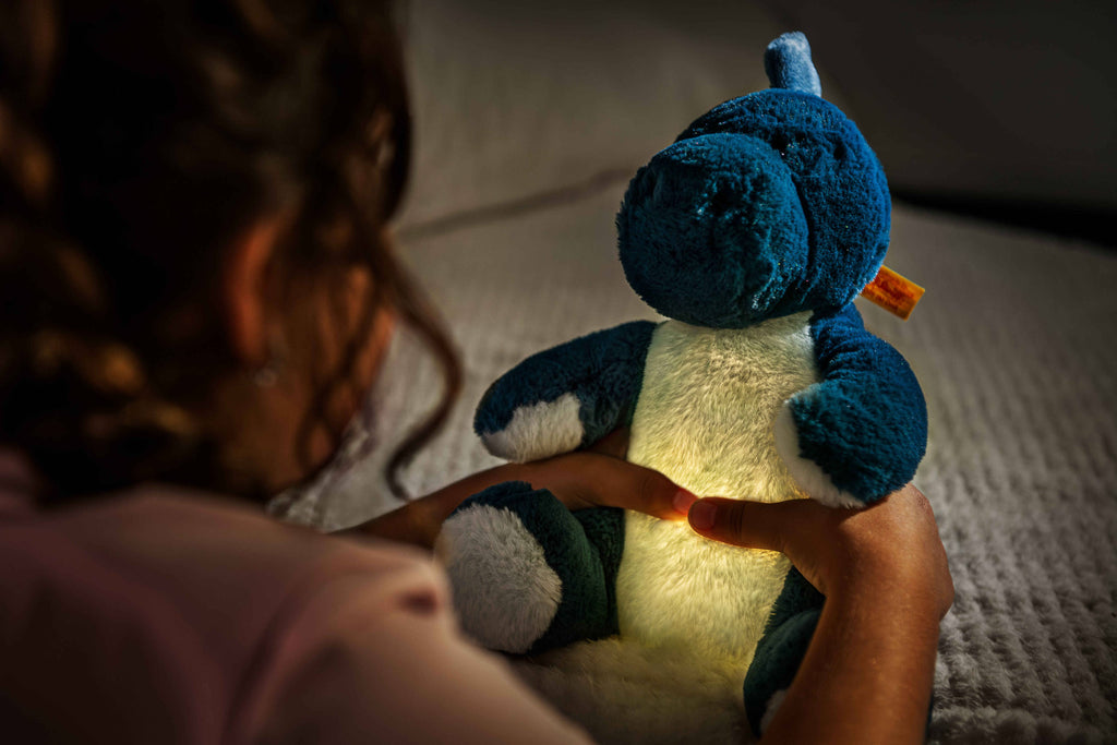 A child sits on a bed in a dimly lit room holding the Steiff Light at Night Spott Stegosaurus Stuffed Plush, 11 inches, which glows softly from its belly.