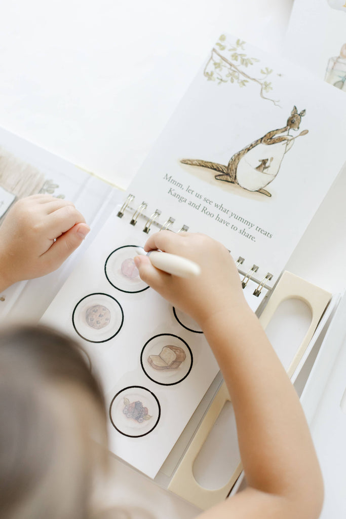 A child uses a water pen to reveal illustrations of food on a page of a Winnie-the-Pooh: Pooh’s Birthday Surprise (Water Reveal) book featuring Kanga and Roo.