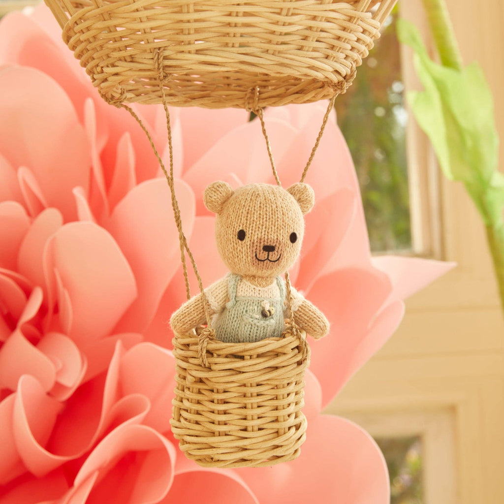 The Cuddle + Kind Tiny Charlie the Honey Bear, hand knit and dressed in light blue overalls, sits in a small woven basket hot air balloon with a large pink paper flower and a window in the background.