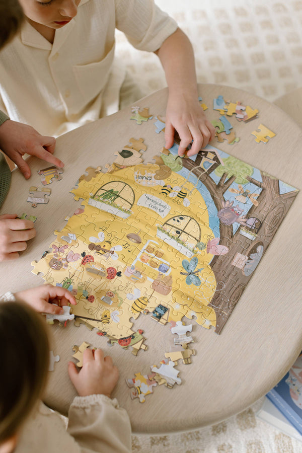 Two children assemble the Honeycomb Cafe 200 Piece Puzzle, featuring a charming house scene with matte textured pieces spread across a light wooden table.