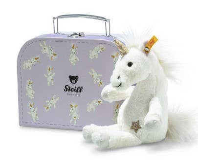 The Steiff Unica Dangling Plush Toy Unicorn, featuring a gold star on its leg, sits in front of a light purple suitcase adorned with unicorn illustrations and a bear logo, promising magical adventures.