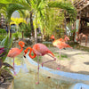 Four Flamingo Finger Puppets stand in shallow water among tropical plants and palm trees, with one balanced on one leg. Their reflections shimmer in a nearby mirror, creating a playful scene perfect for imaginative fun under bright sunlight.