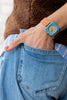 A person wears the Mini Kyomo Watch - Havana Sky, featuring Japanese movement and a tan band, with their hand in the back pocket of blue jeans, dressed in a brown textured top and blue shirt.