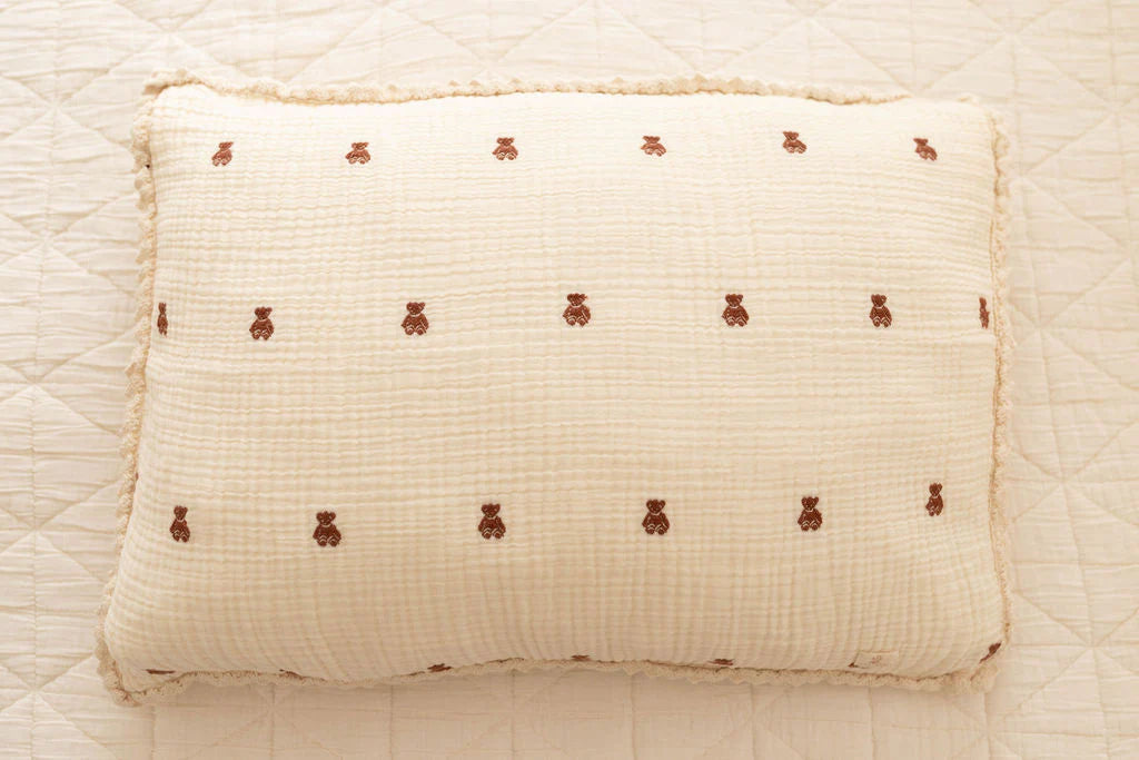 A rectangular pillow with the Organic Cotton Layered Gauze Pillowcase - Teddy, featuring a cream color and small brown teddy bears, rests on a quilted white bedspread.