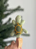 A hand holds the Turtle Finger Puppet, featuring green felt with a yellow shell pattern, artisan-crafted in Nepal. The charming puppet is set against a blurred green tree and white background.