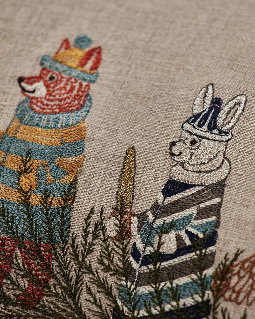 Coral & Tusk Christmas Tree Sled Pillow featuring an embroidered linen design of a bear in a yellow striped sweater and a rabbit in a blue striped sweater standing amongst pine branches.