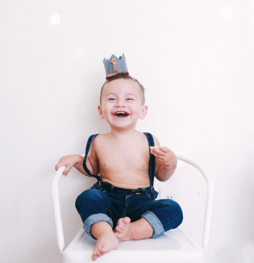 A smiling toddler wearing blue jeans, suspenders, and the Storm Gray Felt Crown sits barefoot on a white chair against a plain white background, ready for birthday celebrations.