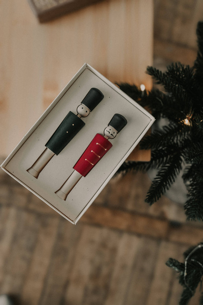 A boxed set of two Wooden Soldiers, nutcracker ornaments in black and red, is placed on a wooden floor. To the right, part of a decorated Christmas tree is visible, evoking the festive spirit similar to opening an advent calendar.