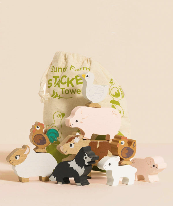 A set of painted wooden farm animal toys, including pig, duck, sheep, goat, rooster, cow, dog, and cat, are arranged before a cloth drawstring bag labeled "Farmyard Stacking Animals and Bag"—an eco-friendly stacking toy.