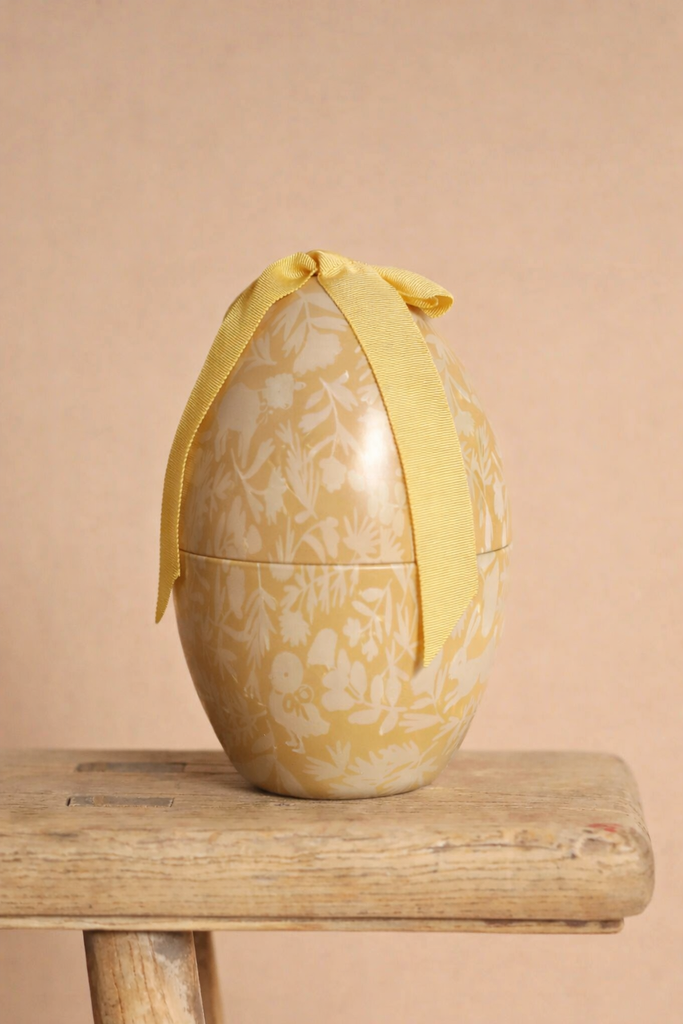 The Maileg Easter Egg, Medium - Cream Yellow, featuring a pale yellow floral pattern and a yellow ribbon bow, sits on a rustic wooden stool against a beige background.