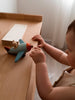 A toddler stands by a wooden table, engaging in imaginative play with the Sabo Concept Aviator Mouse toy airplane.
