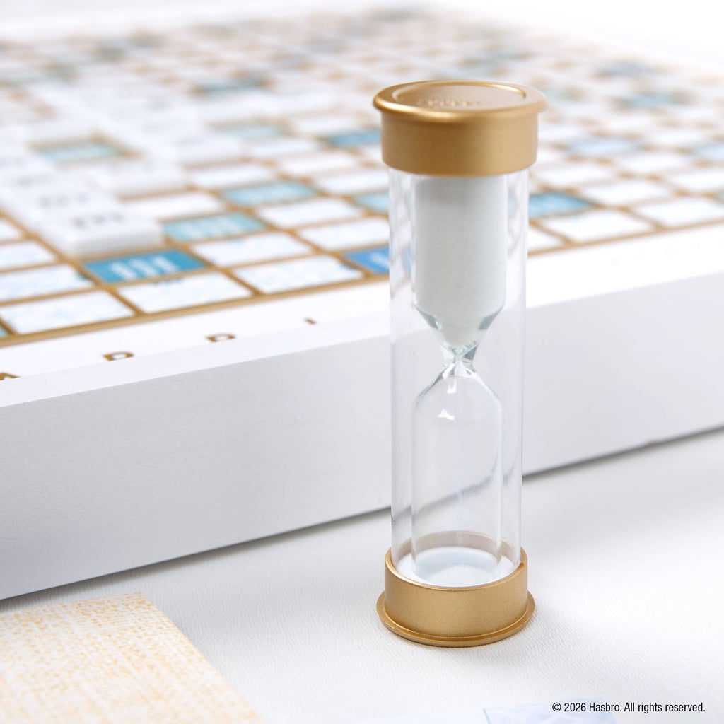 A white hourglass with gold caps stands in the foreground, with a blurred Scrabble Bianco Edition game board in the background.
