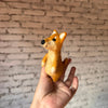 A hand holds a small, handmade Shiba-Inu Dog wool finger puppet in front of a white brick wall background.