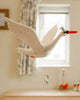 The Handmade Swan Mobile, a plush white swan with a small crown, elegantly flaps as it hangs. Made from organic felt and eco-friendly dyes, it features two shiny bells. Hanging in front of a window, it captures streaming light to softly illuminate the room.