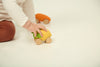 A child in brown pants and a white shirt plays on the floor, pushing the eco-friendly Lou The Lemon Baby Car with green accents. Another orange sensory play car rests in the background on a white surface.