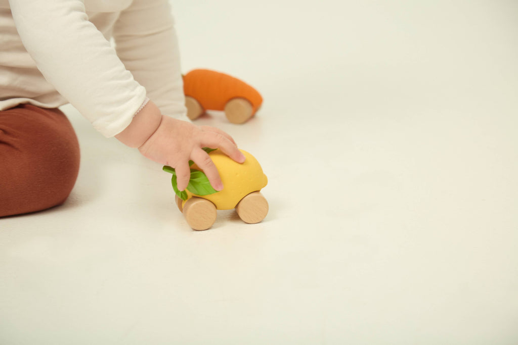 A child in brown pants and a white shirt plays on the floor, pushing the eco-friendly Lou The Lemon Baby Car with green accents. Another orange sensory play car rests in the background on a white surface.