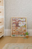 The Beneath the Branches 200 Piece Puzzle, framed and featuring cute animals in a treehouse, leans against floral wallpaper beside a potted plant, a toy dinosaur, and a dresser on a wooden floor.