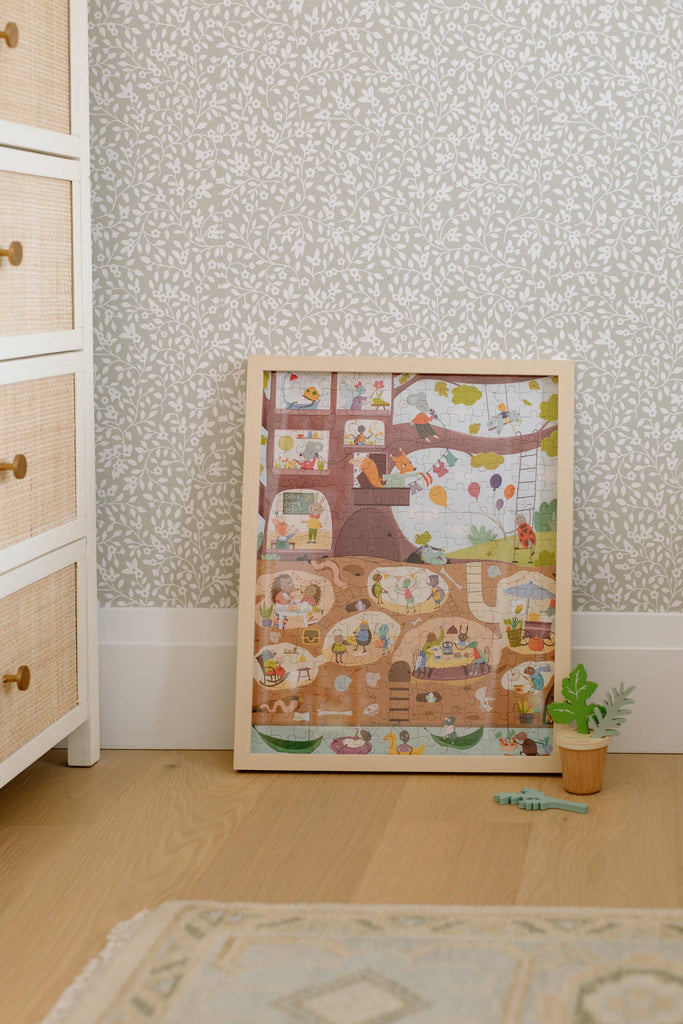 The Beneath the Branches 200 Piece Puzzle, framed and featuring cute animals in a treehouse, leans against floral wallpaper beside a potted plant, a toy dinosaur, and a dresser on a wooden floor.