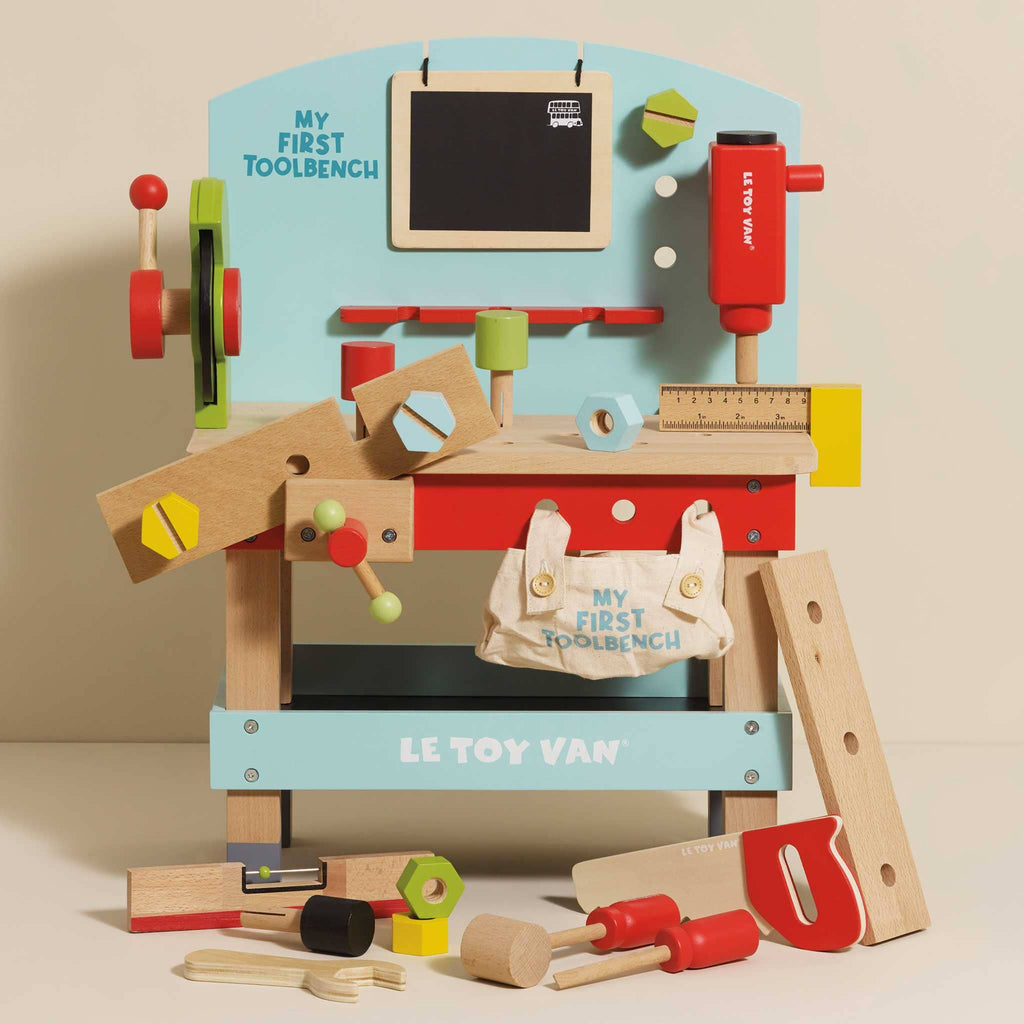 The Wooden Tool Bench is a colorful pretend playset with toy tools—hammer, saw, screwdriver, ruler, nuts, bolts—and a canvas bag labeled “My First Toolbench.” The bench features the “Le Toy Van” brand.