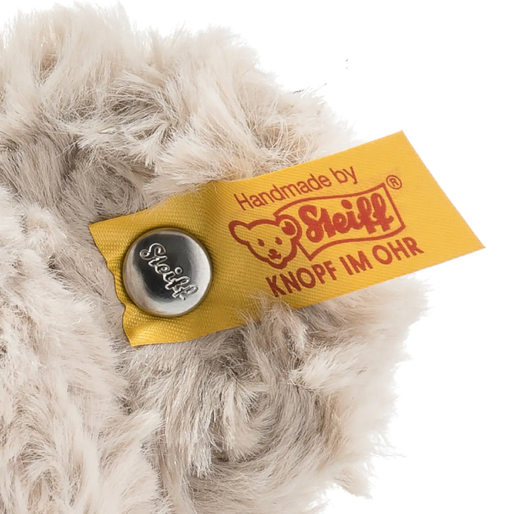 Close-up of a yellow Steiff label on plush fur with text "handmade by Steiff" and the logo, secured with a metal Button in Ear embossed with "Steiff" on it.