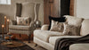 A beige living room features a striped armchair, embroidered pillows, a Coral & Tusk Graveyard Pocket Pillow, lit candles on a wooden table, pumpkins, a Ouija board, and a gray knit blanket on the couch.