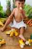 A young child sits on a wooden floor surrounded by bananas, pineapples, lettuce, and carrots, enjoying sensory play with the Ana Banana Teether while eating a banana.