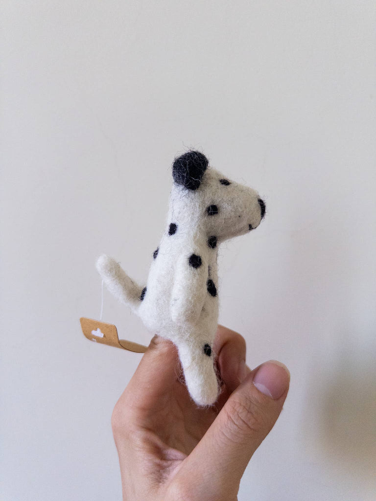A hand holds the Felt Finger Puppet: Dalmatian—a small, felted black-spotted dog with a tag—ideal for imaginative play, against a plain, light background.
