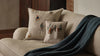 The Coral & Tusk Winter Foxes Pocket Pillow with embroidered fox and tree designs sits on a beige sofa beside a dark green knitted throw, creating a cozy and inviting atmosphere.