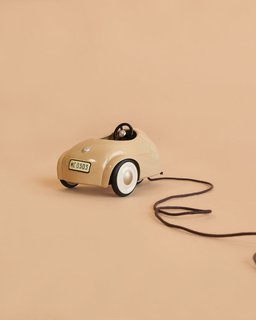 A Maileg Little Sister Driver Set, featuring a small beige car with a string attached, is displayed on a matching beige background. Designed for the Maileg Driver Mouse, the car includes white wheels, a black steering wheel, and a license plate labeled “MC 0303.”.