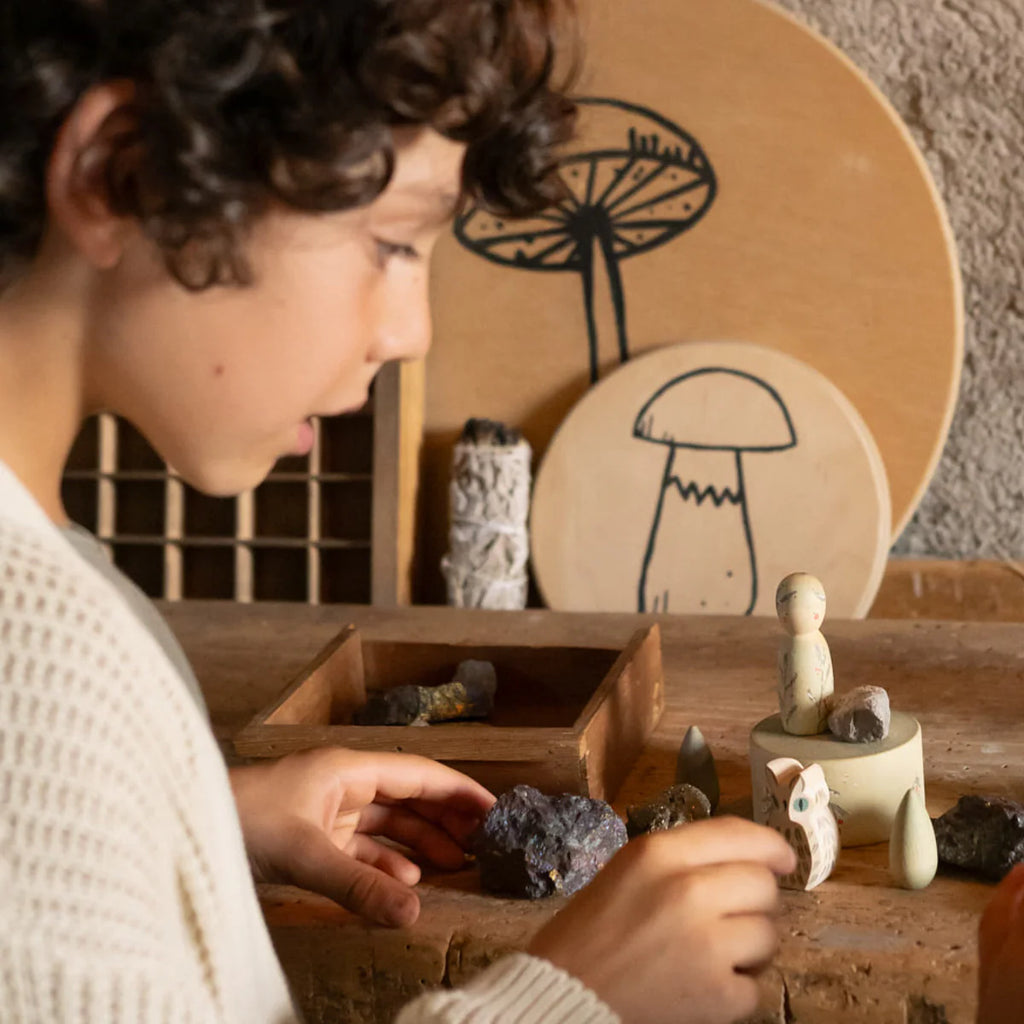 A child with curly hair plays with small rocks and the Hello Winter Set (Grapat X Ostheimer Limited Edition) on a wooden table, surrounded by mushroom art and natural objects.