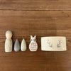 The Hello Winter Set (Grapat X Ostheimer Limited Edition) features five painted wooden toys—a figure, two teardrops, an owl, and a round container—adorned with floral or leaf motifs on a wooden surface.