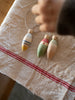 The Grapat Ornament Set - Winter Sparkles features three hand-painted wooden bobbers with string, displayed on a cream cloth with red stripes. A child's hand reaches for these charming ornaments, perfect for magical home decor or festive decoration.