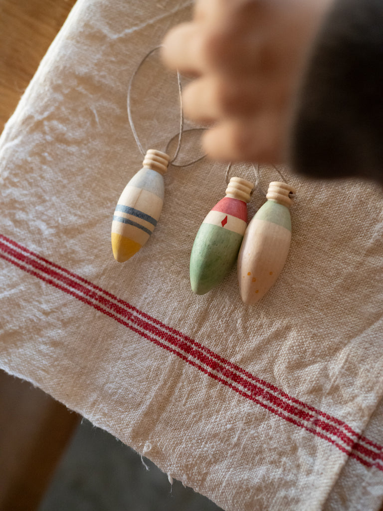 The Grapat Ornament Set - Winter Sparkles features three hand-painted wooden bobbers with string, displayed on a cream cloth with red stripes. A child's hand reaches for these charming ornaments, perfect for magical home decor or festive decoration.